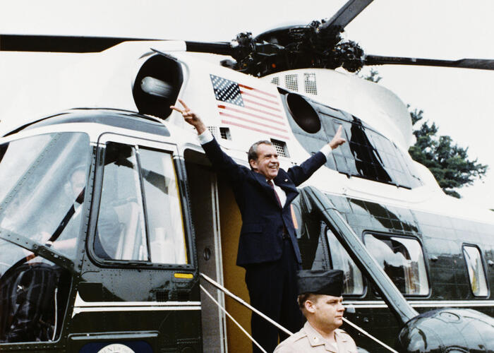 Military man standing guard in front of a helicopter as the President raises hands in victory sign.