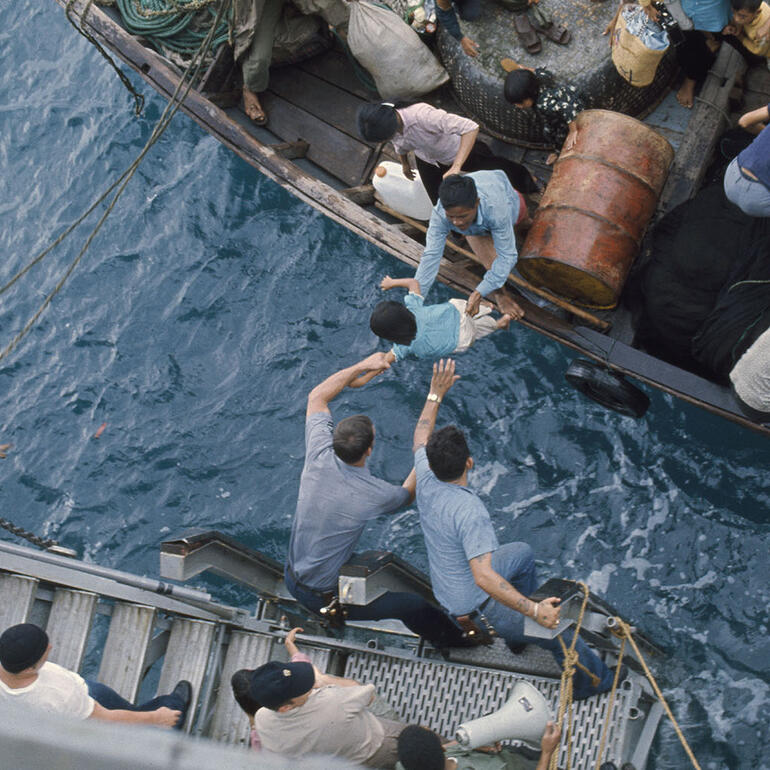Color photograph of crewmen helping refugees on a craft in the water