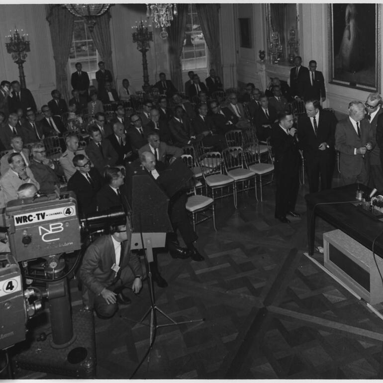 black and white photograph of President signing resolution in front of an audience and media cameras and 