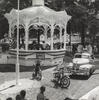 Black and white photograph of town square with bunting, vehicles, and boys riding bicycles