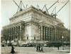Photograph of building under construction with people, cars, and trolley in front.