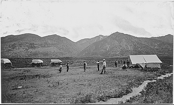 Black and white image of men standing in front of tents  on a plain in front of a mountain range.