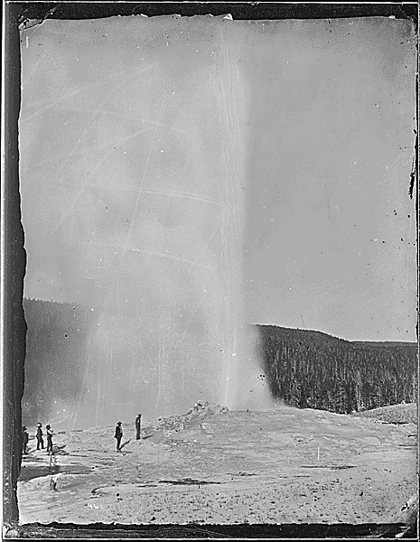 Black and white image of geyser erupting with people standing in the foreground.