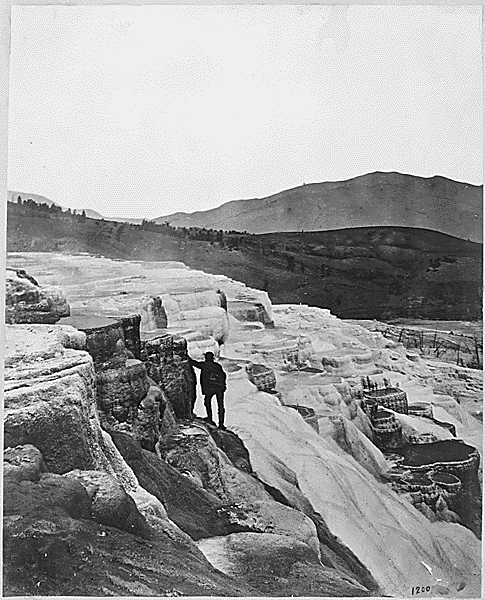 Black and white image of man standing next to rocks near hot springs.