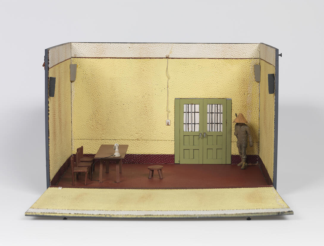 Color photograph of small model room with interrogation table, green doors, and man standing in corner