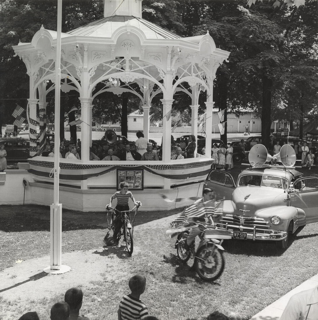 Black and white photograph of town square with bunting, vehicles, and boys riding bicycles
