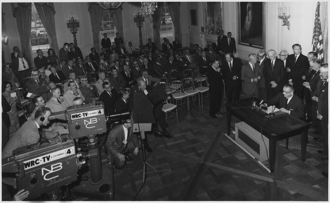 black and white photograph of President signing resolution in front of an audience and media cameras and 