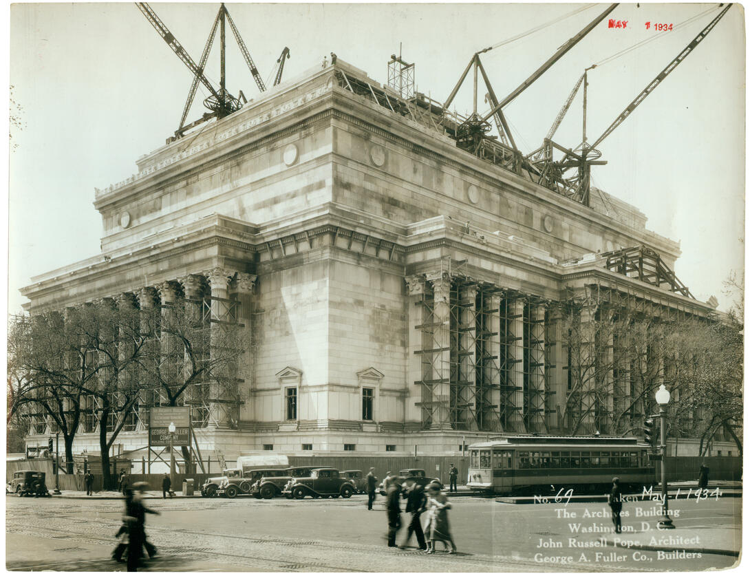 Photograph of building under construction with people, cars, and trolley in front.