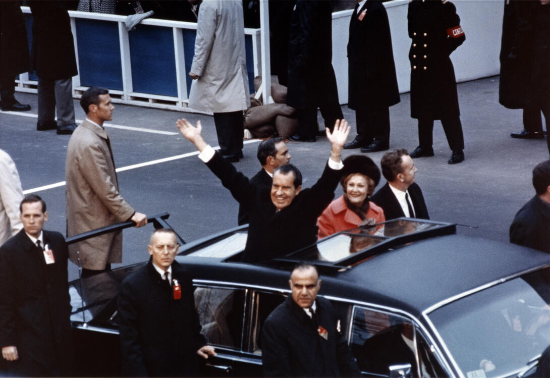 Color image of president waving from motorcade surrounded by secret service agents